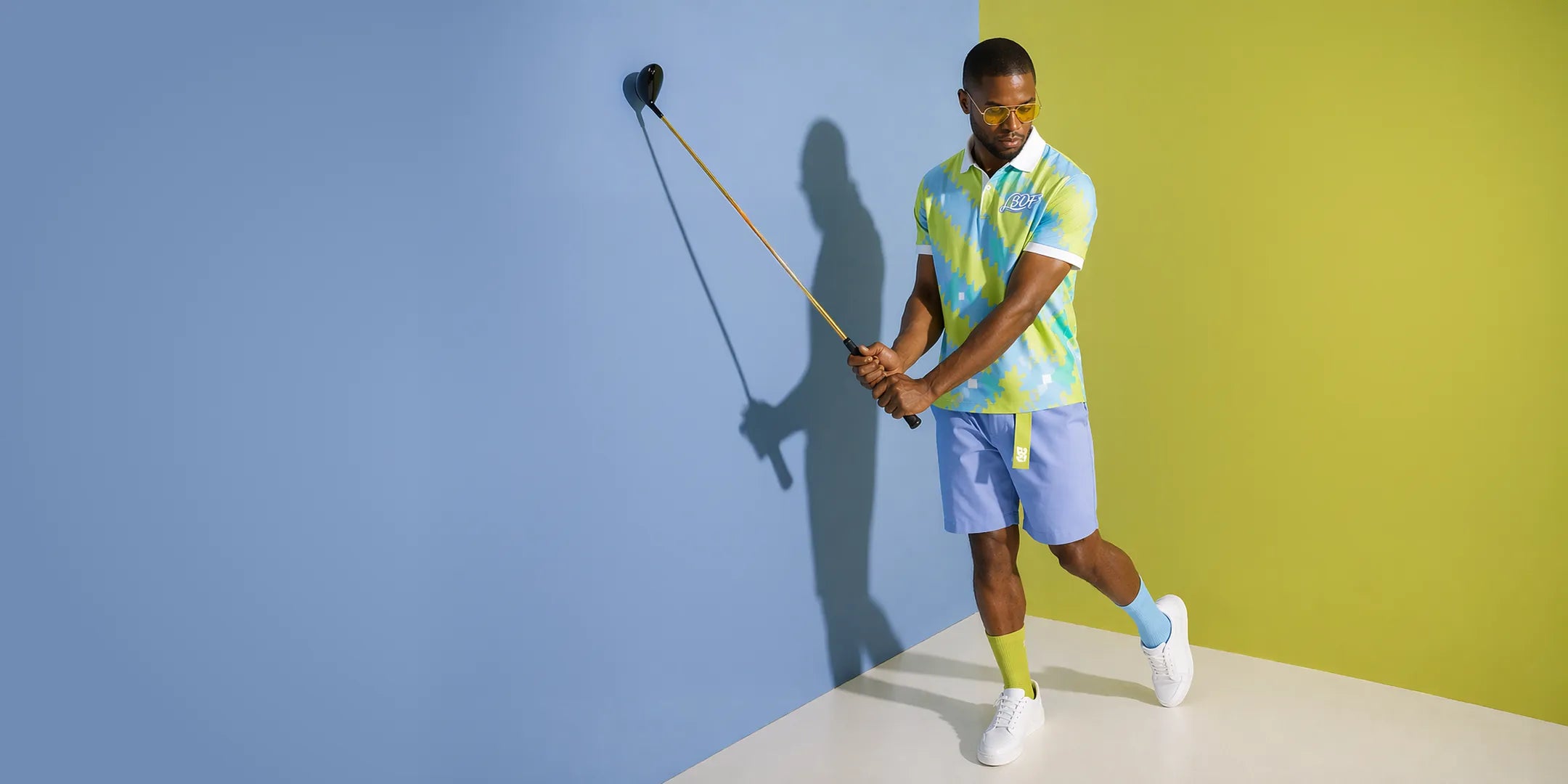 Man in colorful shirt and shorts holding a golf club against a multicolored wall.